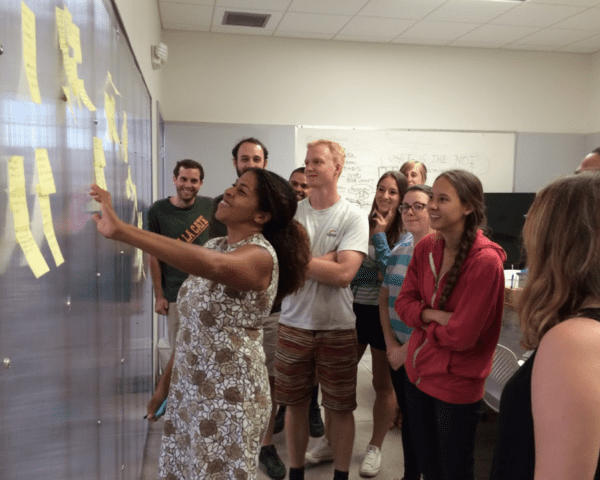 screen_shot_2017-01-30_at_12 Albert and Tina Small Center for Collaborative Design A group of people standing in front of a wall covered with sticky notes. One person is pointing at the notes while others watch attentively. The setting appears to be a classroom or meeting room, with a whiteboard visible in the background