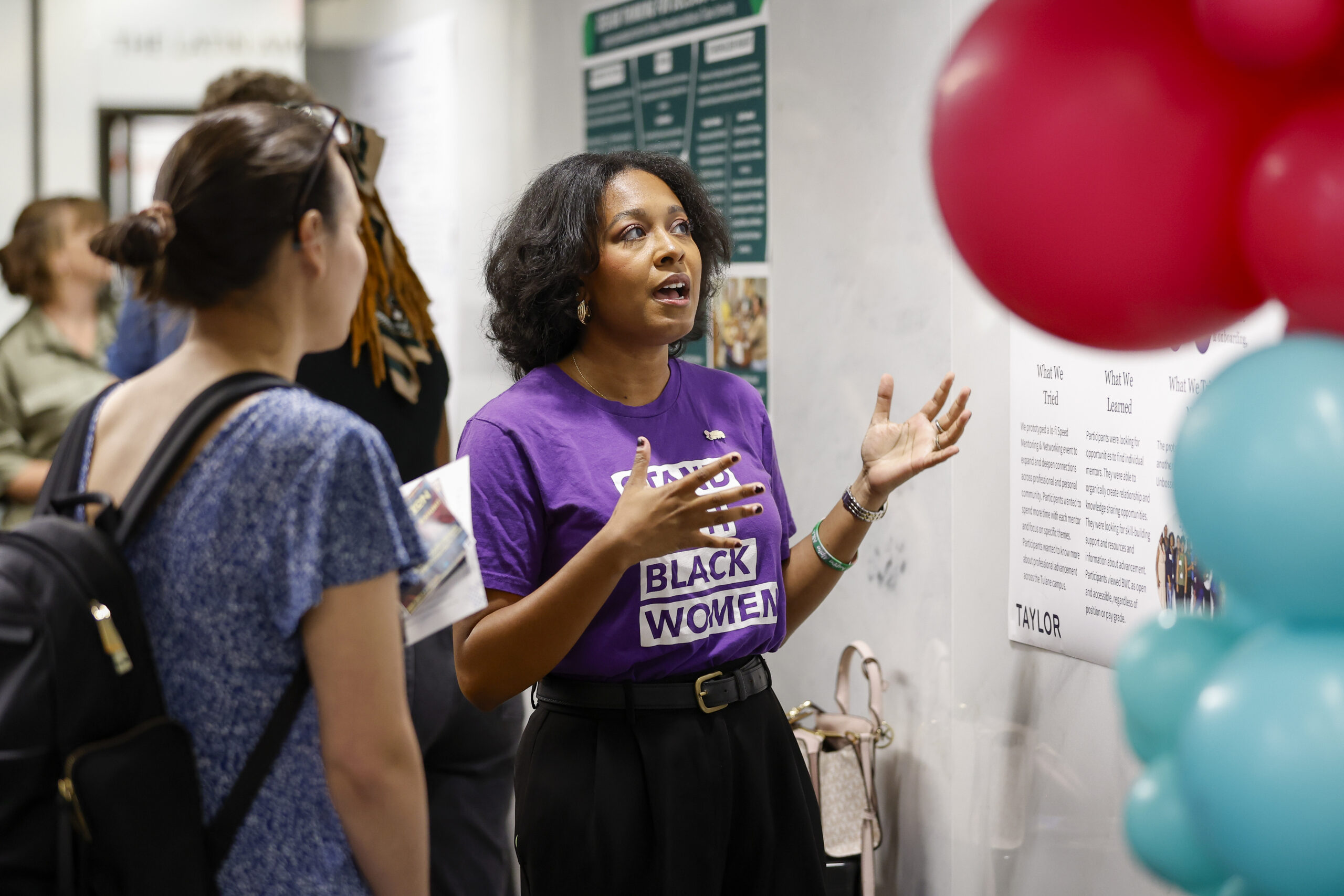 Taylor 10: Homecoming Highlights A woman in a purple "Centering Black Women" t-shirt is passionately speaking and gesturing with her hands while presenting research or a project to another woman. Behind them are posters on the wall labeled “What We Tried,” “What We Learned,” and “TAYLOR.” Pink and teal balloons are partially visible in the foreground.