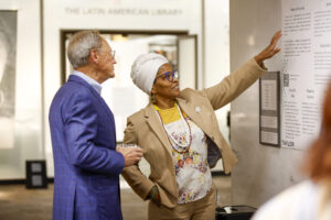 Taylor 10: Homecoming Highlights A woman in a beige suit and white headwrap gestures toward an informational poster while speaking to a man in a blue checkered suit. They are standing in a well-lit exhibition space near a sign that reads “The Latin American Library.”
