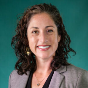 A professional headshot of Julia Lang with a confident expression, wearing a black top and a grey blazer. She stands against a green background, looking directly at the camera with a poised demeanor.