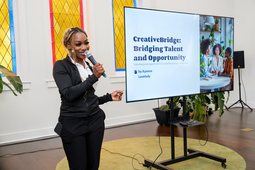 ChangemakerApril2025-122 Ayanna Lovelady stands smiling with a microphone in hand during a presentation. She is dressed in a black and white outfit and stands next to a large screen displaying her project titled "CreativeBridge: Bridging Talent and Opportunity," with a subtitle about unlocking paid opportunities for talented young professionals. The background includes vibrant stained glass windows and large green plants, suggesting a warm and inspiring event setting.
