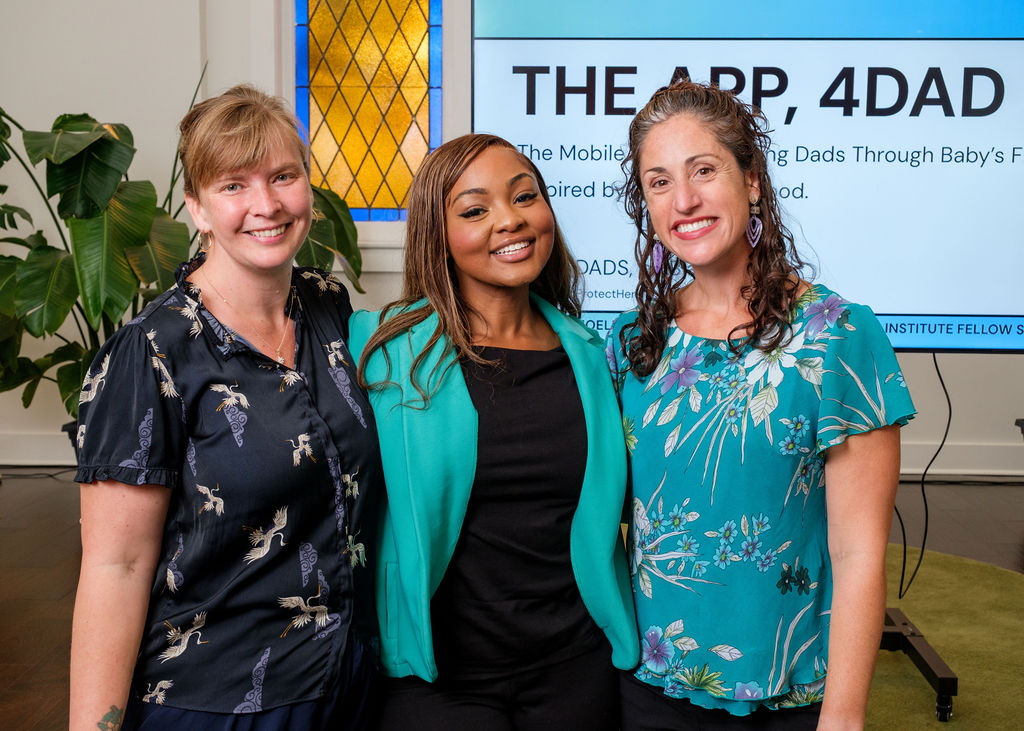ChangemakerApril2025-140 Three women stand smiling together in front of a screen that reads “THE APP, 4DAD.” The woman on the left wears a dark blouse with a crane print, the woman in the center wears a turquoise blazer over a black outfit, and the woman on the right wears a floral teal blouse. A large leafy plant and stained-glass window are visible in the background.