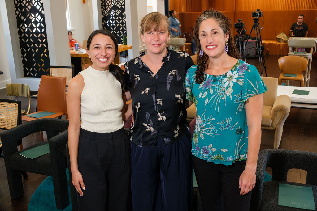 ChangemakerApril2025-7 Three women stand close together, smiling warmly at the camera in a well-lit room with modern furniture and a camera setup visible in the background. On the right is Julia Lang, wearing a teal floral top and purple earrings. In the center, a woman with blonde hair in a ponytail wears a navy blouse with a crane bird pattern and matching navy pants. On the left, a woman with long dark hair in a low ponytail wears a sleeveless ivory top paired with high-waisted black pants.